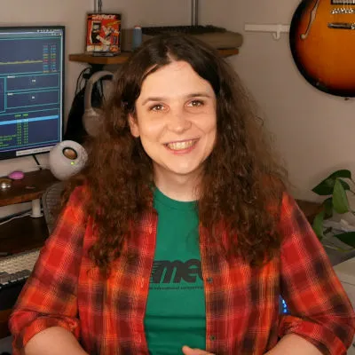 A photo of me (a mid-30s white lady with brown curly hair). I'm smiling at the camera and wearing a red plaid shirt with a green MECC shirt under it. I'm sitting at a desk with a computer over one shoulder and a guitar over the other.