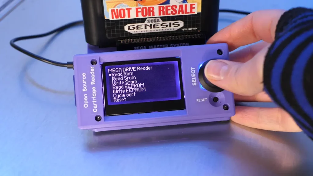 Sonic the Hedgehog (for Sega) is in a purple contraption which says "Open Source Cartridge Reader" and my thumb is pressing a large knob/button on it. A screen on the contraption says "MEGA DRIVE Reader" and the option "Read Rom" is selected.